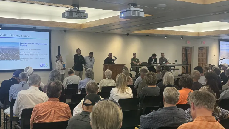 A community meeting about a solar and battery storage project, with attendees seated in a conference room. A panel of speakers stands near a presentation screen displaying project details. The audience listens attentively as speakers address questions.