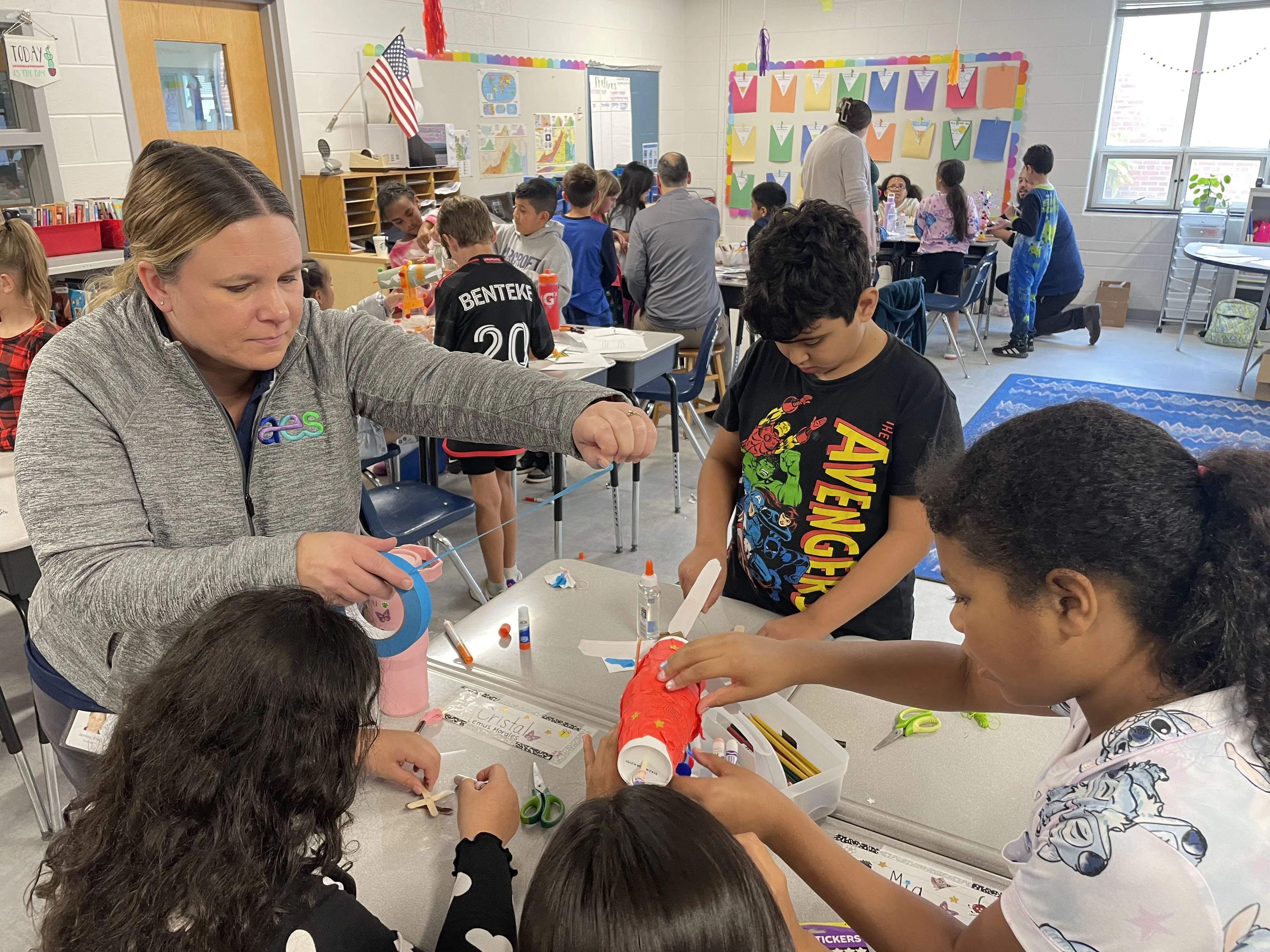 A teacher wearing a gray jacket with an AES logo assists a group of elementary school students during a hands-on craft activity in a classroom. The teacher holds a roll of blue tape, helping the students assemble their project. The children are engaged, using colorful materials like glue, scissors, and construction paper to create their crafts