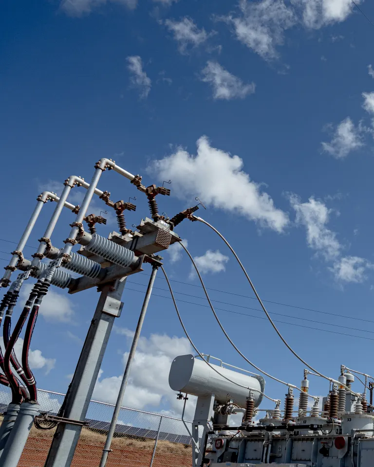 An electrical substation with various transformers, insulators, and high-voltage power lines set against a bright blue sky with scattered clouds. The infrastructure includes metal structures supporting electrical components, illustrating power distribution technology.