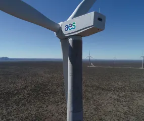 Close-up of a large wind turbine with the AES logo on its side, set in a vast open landscape with several other wind turbines in the distance under a clear blue sky.
